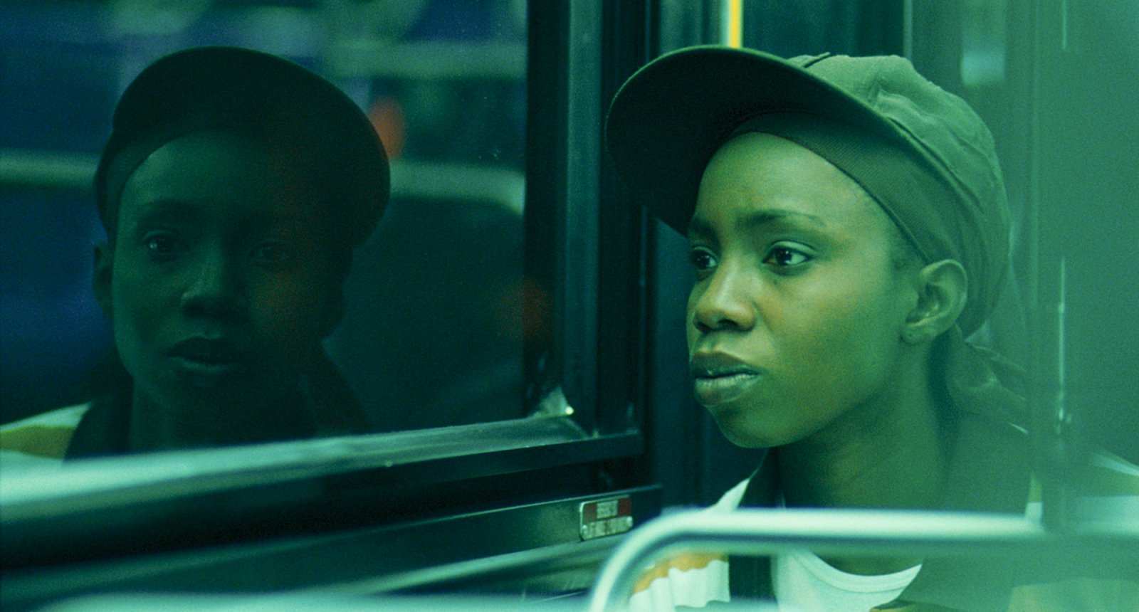 A Black woman in a baseball hat over a brown durag is sitting on a bus. She is looking seriously out the window, and her reflection in the bus window is looking at the viewer of the image. The overall lighting of the image is green, with additional texture on the right side of the image.